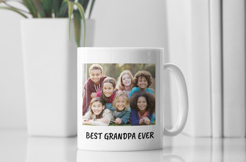 Op de afbeelding: Witte keramische mok met een foto van zeven kinderen en de tekst "BEST GRANDPA EVER" in het zwart. De kinderen lachen en liggen op het gras. De mok heeft een wit oor en staat op een wit oppervlak.