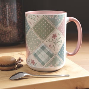 May include: A large ceramic mug with a patchwork design in pastel colors. The mug features squares with polka dots, floral patterns, and plaid. The handle and interior rim are a soft pink. A spoon and a macaron are on a wooden surface.