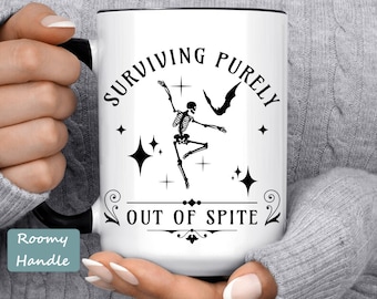 May include: White ceramic mug with a black rim and handle. The mug features a black and white illustration of a skeleton dancing with a bat and stars. The text "SURVIVING PURELY OUT OF SPITE" is printed in black on the mug.