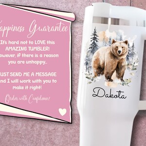 Pode incluir: Caneca branca com uma ilustra&ccedil;&atilde;o em aquarela de um urso pardo em um cen&aacute;rio de floresta, com o nome "Dakota" em escrita cursiva. Um pergaminho rosa com o texto "Happiness Guarantee" est&aacute; no fundo.