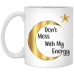 Puede incluir: Taza de cer&aacute;mica blanca con un dise&ntilde;o de media luna y estrella dorada. La taza presenta el texto "Don't Mess With My Energy" en negro. El asa es blanca.