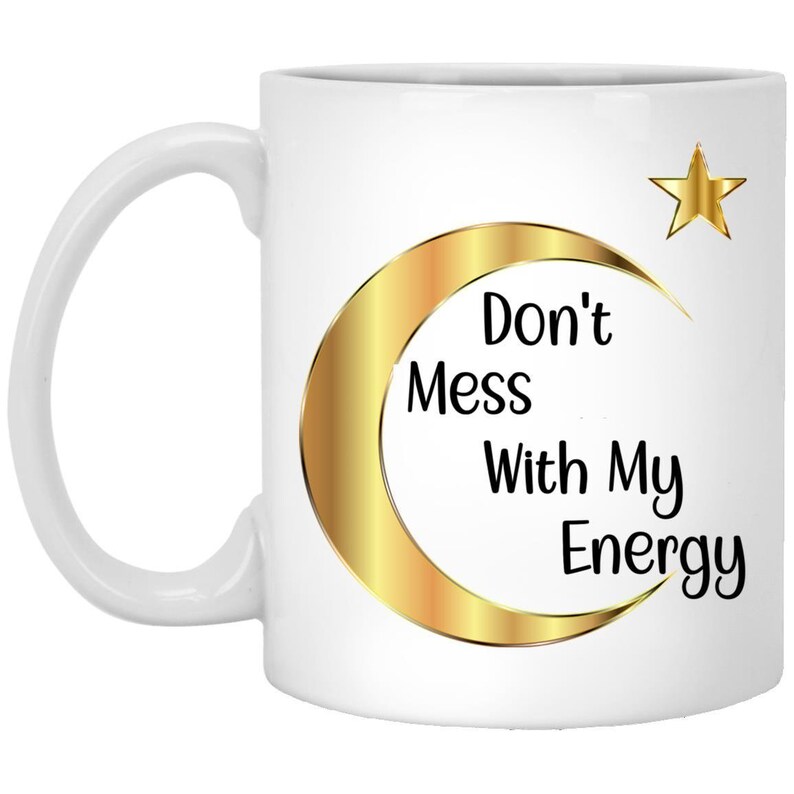 Puede incluir: Taza de cer&aacute;mica blanca con un dise&ntilde;o de media luna y estrella dorada. La taza presenta el texto "Don't Mess With My Energy" en negro. El asa es blanca.