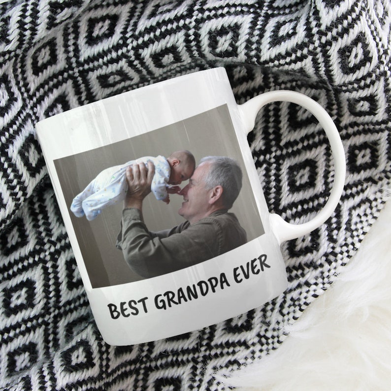 Op de afbeelding: Witte keramische mok met een foto van een opa en een baby. De mok heeft de tekst "BEST GRANDPA EVER". De mok staat op een zwart-wit patroon deken.