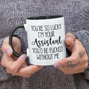 May include: White ceramic mug with a black interior and handle. The mug features the text "YOU'RE SO LUCKY I'M YOUR Assistant YOU'D BE FUCKED WITHOUT ME" in black lettering. The mug is held by a person.