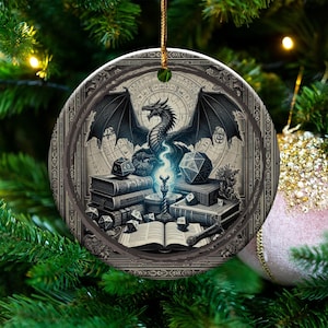 May include: Round Christmas ornament featuring a detailed illustration of a dragon with spread wings, books, and dice. The ornament has a dark, vintage aesthetic with a detailed border and is suspended by a gold string. The background is a Christmas tree.