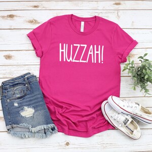 May include: A pink t-shirt with the word "HUZZAH!" printed in white on the front. The shirt is paired with denim shorts and white sneakers.