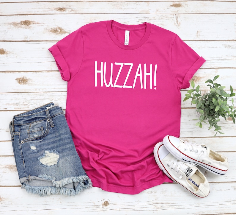 May include: A pink t-shirt with the word "HUZZAH!" printed in white on the front. The shirt is paired with denim shorts and white sneakers.