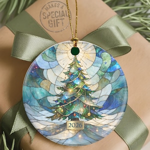 May include: A round, flat Christmas tree ornament featuring a stained-glass style illustration of a decorated evergreen tree in shades of green and blue. The design includes glowing lights, a star at the top, and the year 2026 printed at the base.