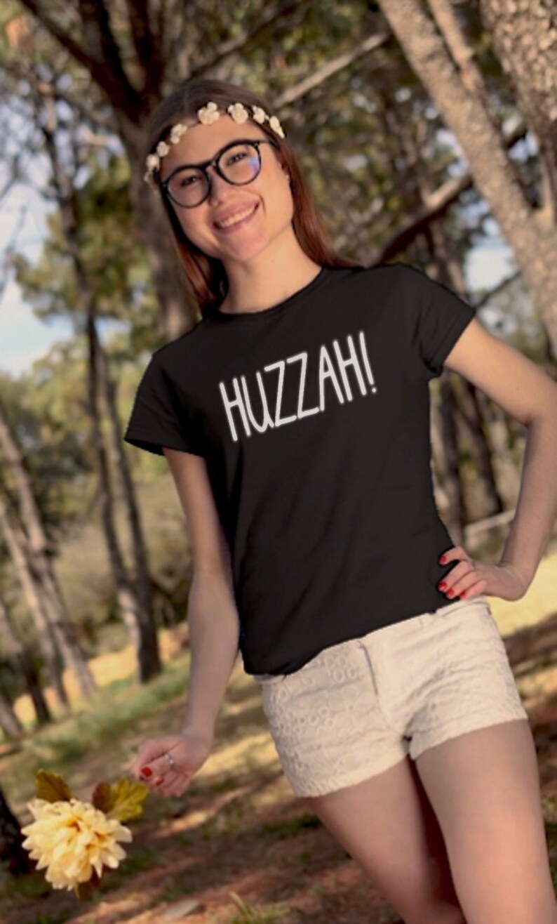 May include: A black t-shirt with white text that says "HUZZAH!" The person wearing the shirt is standing in a forest and holding a yellow flower.