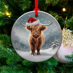 May include: A round Christmas ornament featuring a Highland cow wearing a Santa hat. The cow is brown with long horns, set against a snowy mountain scene. The ornament has a red ribbon for hanging.
