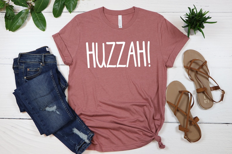 May include: A mauve t-shirt with the word "HUZZAH!" printed in white on the front.