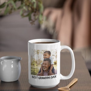 Op de afbeelding: Witte keramische mok met een foto van een opa en kleinkind, met de tekst "BEST GRANDPA EVER". De mok staat op een houten tafel met een kleine witte kan en een houten lepel.