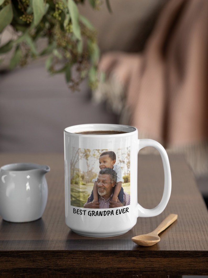 Op de afbeelding: Witte keramische mok met een foto van een opa en kleinkind, met de tekst "BEST GRANDPA EVER". De mok staat op een houten tafel met een kleine witte kan en een houten lepel.