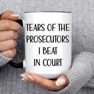 May include: White ceramic mug with a black handle and rim. The mug features the text "TEARS OF THE PROSECUTORS I BEAT IN COURT" in black, bold letters. The mug is held by a person wearing a gray sweater.