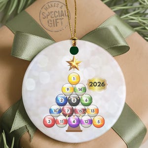 May include: A round white ceramic ornament featuring a graphic of billiard balls arranged in a triangular Christmas tree shape, topped with a gold star. The design includes the year 2026 in a gold-bordered label and hangs from a gold string.