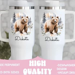 Pode incluir: Caneca de viagem branca com uma al&ccedil;a grande, com uma ilustra&ccedil;&atilde;o em aquarela de um urso pardo em uma cena de floresta. O nome "Dakota" est&aacute; escrito abaixo. A caneca tem tampa e al&ccedil;a brancas.