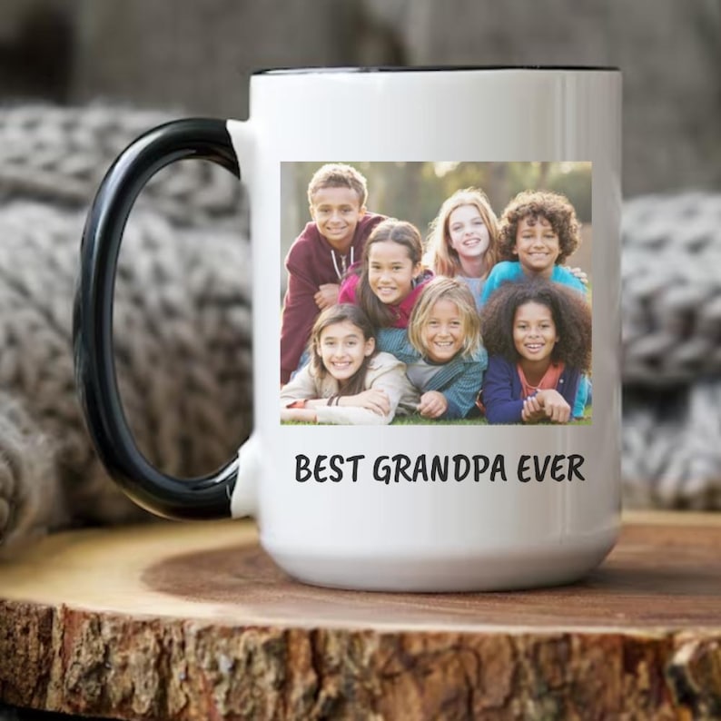 Op de afbeelding: Witte keramische mok met een zwarte handgreep en rand. De mok heeft een foto van kinderen en de tekst "BEST GRANDPA EVER". De mok staat op een houten oppervlak.