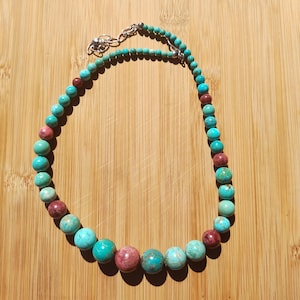 May include: A necklace made of turquoise and maroon beads, with a silver clasp. The beads are of varying sizes and are arranged in a graduated pattern. The necklace is presented on a wooden surface, showcasing its colours.