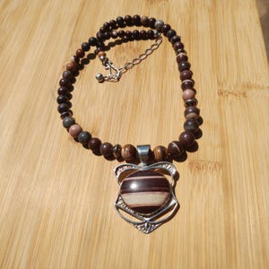 May include: A beaded necklace featuring a pendant with a striped, oval-shaped stone in a silver-colored setting. The necklace has a chain and is composed of various brown and tan beads. The necklace is displayed on a wooden surface.
