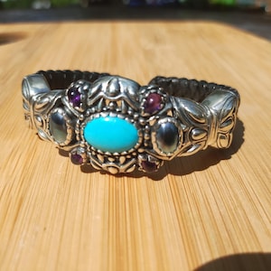 May include: Silver bracelet featuring a central turquoise oval stone, accented by smaller purple and grey gemstones. The bracelet has an intricate design and a black, braided band. The jewellery is presented on a wooden surface.