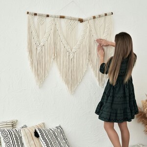 May include: A white macrame wall hanging with a wooden dowel rod. The wall hanging is made of knotted cords and has a fringe at the bottom. A person is adjusting the wall hanging on a white wall.