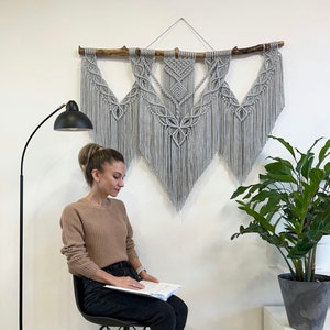 May include: A grey macrame wall hanging with a wooden branch at the top. The wall hanging is made of knotted cords and has a fringe at the bottom. The wall hanging is hanging on a white wall.