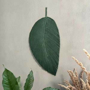 May include: A green macrame leaf wall hanging with a fringed edge. The leaf is hanging on a light green wall.
