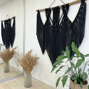 May include: A large, black macrame wall hanging with a wooden dowel. The intricate design features knotted patterns and long fringe. The wall decor is displayed on a white wall, with a mirror reflecting the design.