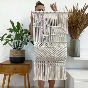 May include: White macrame wall hanging with a pocket and fringe. The wall hanging features a geometric design and is made with natural cotton cord.
