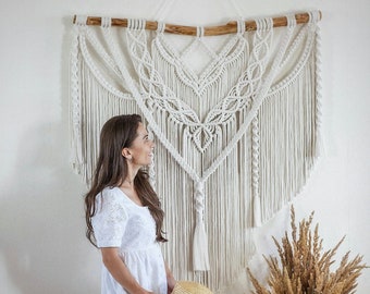 May include: A white macrame wall hanging with a wooden dowel at the top. The wall hanging is in the shape of a heart and has a fringe at the bottom. A woman is sitting on a wooden stool in front of the wall hanging, holding a straw hat.