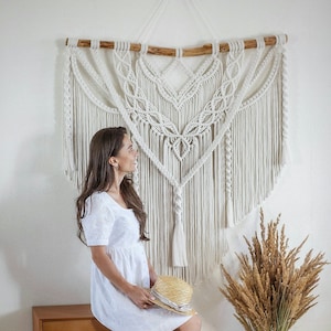 Puede incluir: Un tapiz de macramé blanco con una barra de madera en la parte superior. El tapiz tiene forma de corazón y un fleco en la parte inferior. Una mujer está sentada en un taburete de madera frente al tapiz, sosteniendo un sombrero de paja.