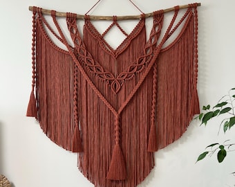 May include: A large, rust-colored macrame wall hanging with a wooden branch at the top. The macrame is made with a variety of knots and has a geometric design. The hanging has a fringe at the bottom.
