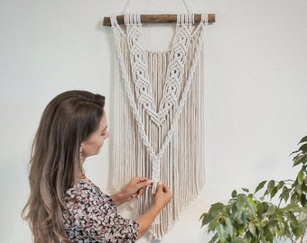 May include: A woman in a floral dress hangs a white macrame wall hanging on a white wall. The wall hanging is made of cotton cord and has a wooden dowel at the top.