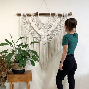Könnte beinhalten: Ein großer, weißer Makramee-Wandbehang mit geometrischem Muster. Der Behang ist aus Baumwollschnur gefertigt und an einer Holzleiste befestigt. Der Behang wird an einer weißen Wand mit einer Topfpflanze und einem Holz-Beistelltisch im Vordergrund gezeigt.