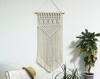 May include: A white macrame wall hanging with a geometric pattern, hanging on a white wall. There is a white chair with a straw bag and a hat on it. There are two green plants in pots on the right side of the image.