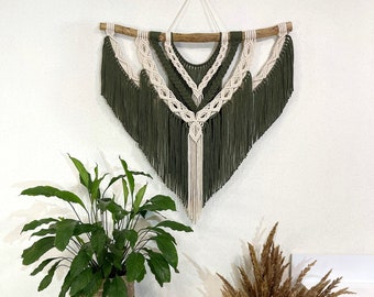 May include: A green and white macrame wall hanging with fringe, featuring a geometric design. The hanging is displayed on a white wall with a potted plant and a vase of dried pampas grass.
