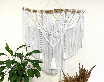 May include: White macrame wall hanging with a wooden branch at the top and a geometric pattern. The hanging is displayed on a white wall with a wooden side table and a vase with dried flowers.