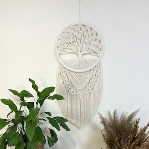 Op de afbeelding: Een witte macramé wandhanger met een levensboomontwerp. De hanger is rond met een franje van wit garen. De hanger wordt getoond op een witte muur met een potplant en gedroogd pampasgras.