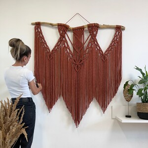 May include: A large, rust-coloured macrame wall hanging with a wooden branch at the top. The hanging features a geometric pattern and long fringe.