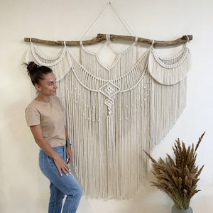 Op de afbeelding: Grote macramé wandhanger in crèmekleur, opgehangen aan een natuurlijke drijfhouttak. Het ingewikkelde ontwerp heeft geknoopte patronen en franjes, waardoor een bohemien esthetiek ontstaat. Een decoratief stuk voor de interieurdecoratie.