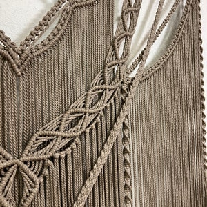 May include: A beige macrame wall hanging with a geometric pattern. The hanging is made of thick rope and has a natural wood dowel at the top.