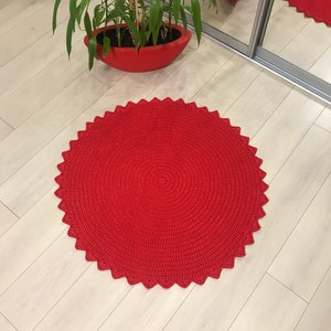 May include: A red crocheted round rug with a scalloped edge. The rug is on a light wood floor.