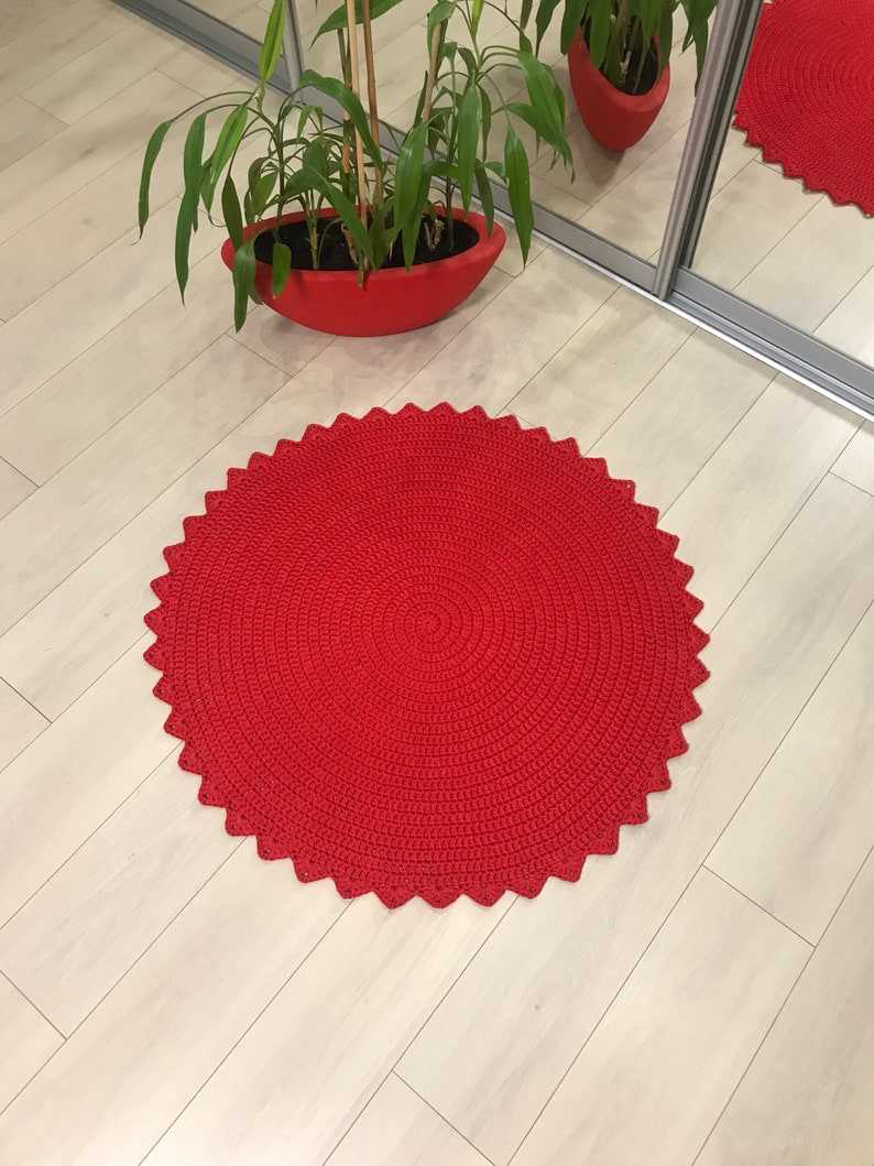 May include: A red crocheted round rug with a scalloped edge. The rug is on a light wood floor.