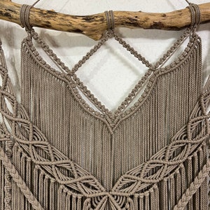 May include: A macrame wall hanging with a natural wood branch at the top. The macrame is made with a light brown cord and features a geometric pattern with fringe.