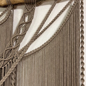 May include: A macrame wall hanging with a natural wood branch at the top. The hanging is made with brown cord and features a variety of knots and patterns.