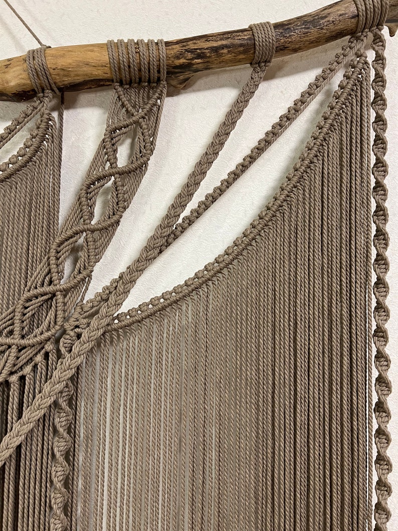 May include: A macrame wall hanging with a natural wood branch at the top. The hanging is made with brown cord and features a variety of knots and patterns.