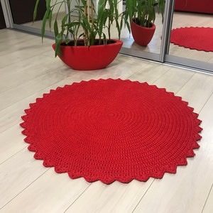 May include: A red crocheted round rug with a scalloped edge. The rug is on a light wood floor.