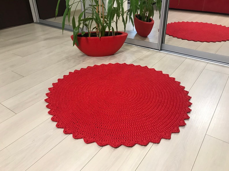 May include: A red crocheted round rug with a scalloped edge. The rug is on a light wood floor.