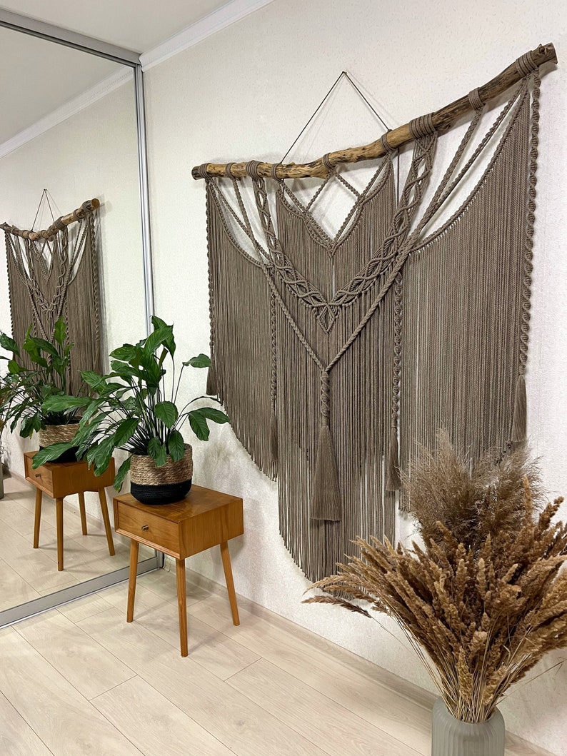 May include: A large macrame wall hanging with a natural wood branch at the top. The macrame is made with a light brown cord and has a geometric design. The wall hanging is hanging on a white wall in a room with a wooden floor.