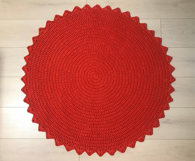 May include: A round, red crocheted rug with a scalloped edge. The rug is made of a thick, textured yarn and has a circular pattern.